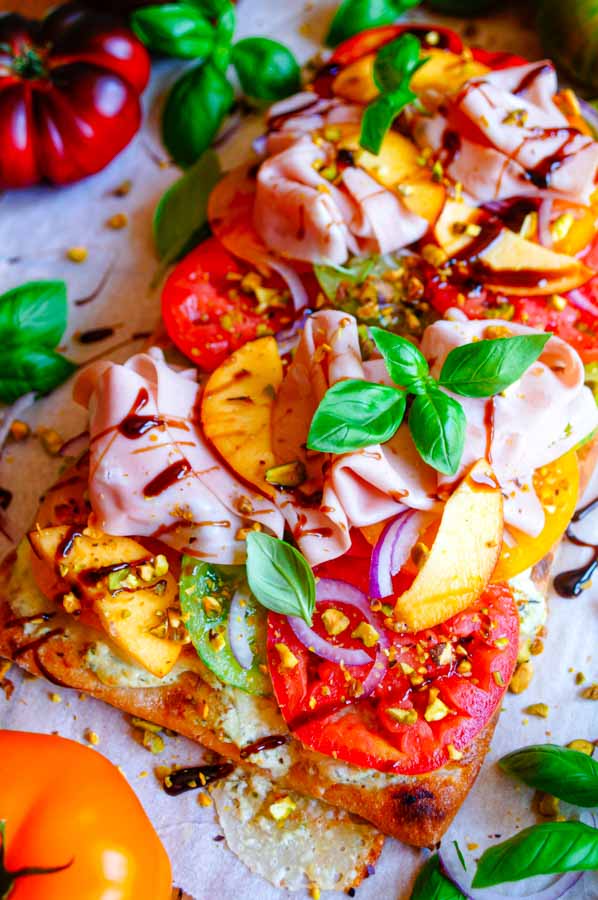 Lekker makkelijk recept voor Pinsa met tomaten en pesto, simpel doordeweekse maaltijd voor op weekmenu of als hapje bij een zomerse apero. Het platbrood wordt belegd met een romig pesto-ricotta crème en mozzarella. Daarop komen heerlijke volle tomaten, plakjes perzik en mortadella.