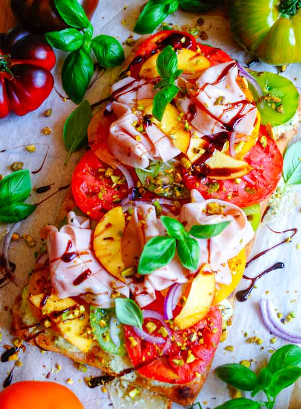 Lekker makkelijk recept voor Pinsa met tomaten en pesto, simpel doordeweekse maaltijd voor op weekmenu of als hapje bij een zomerse apero. Het platbrood wordt belegd met een romig pesto-ricotta crème en mozzarella. Daarop komen heerlijke volle tomaten, plakjes perzik en mortadella.
