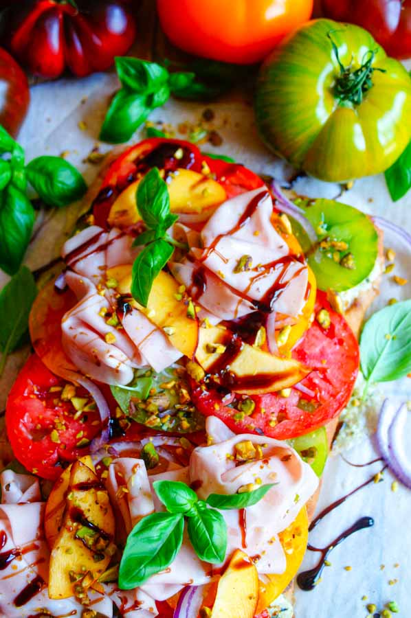 Lekker makkelijk recept voor Pinsa met tomaten en pesto, simpel doordeweekse maaltijd voor op weekmenu of als hapje bij een zomerse apero. Het platbrood wordt belegd met een romig pesto-ricotta crème en mozzarella. Daarop komen heerlijke volle tomaten, plakjes perzik en mortadella.